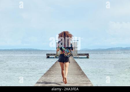 Rückansicht einer unkenntlich frauen Frau, die auf einem schäbigen Holzpier mit Toren läuft, die die Freiheit auf dem Meer genießen Stockfoto