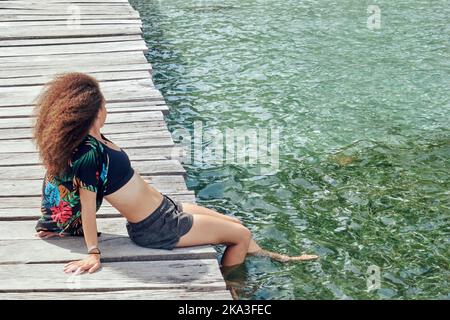 Ganzkörper-Seitenansicht einer ethnischen Frau mit lockigen Haaren in Shorts mit Oberteil und Hemd auf einem schäbigen Holzsteg mit Beinen in klarem blauem Wasser Stockfoto