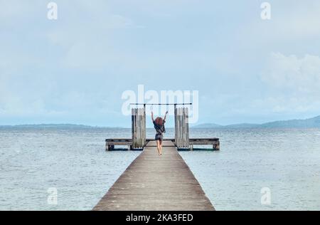 Rückansicht der fernen Frau, die auf einem hölzernen schäbigen Pier mit Toren geht und die Arme hochhebt und die Freiheit auf dem Meer genießt Stockfoto