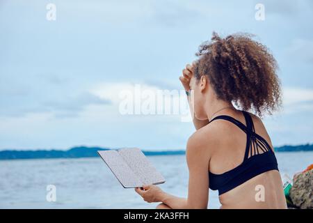 Rückansicht einer fokussierten hispanischen Hündin in schwarzer Spitze, die mit einem Tagebuch am tropischen Strand mit Palmen sitzt und die Kamera anschaut Stockfoto