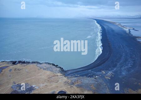 Atemberaubende Drohne Blick auf schwarzen vulkanischen Strand in der Nähe schneebedeckter Berge an der Küste des winkenden Meeres am Abend in Island Stockfoto