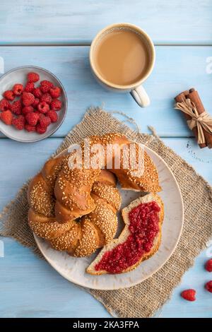 Hausgemachtes süßes Brötchen mit Himbeermarmelade und Tasse Kaffee auf blauem Holzhintergrund und Leinentextilien. Draufsicht, flach liegend, Nahaufnahme. Stockfoto