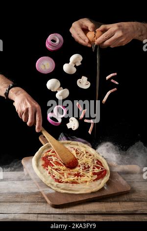 Ernte Hände brechen Ei und verteilen Sauce und Käse mit Spatel Pizza, während Pilze und Zwiebel mit Speck auf schwarzen Hintergrund fallen Stockfoto