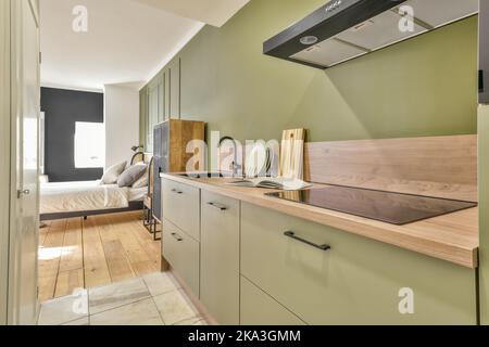 Theke mit Waschbecken und verschiedenen Geschirrs in der modernen Küche mit grünen Schränken und modernen Küchenutensilien neben dem Schlafzimmer in stilvollem, offenem Schnitt platziert Stockfoto