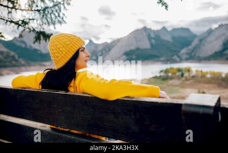 Seitenansicht Frau in orangefarbenem Pullover und Mütze, die auf einer Holzbank sitzt und die atemberaubende Landschaft von Bergrücken und ruhigem See genießt Stockfoto