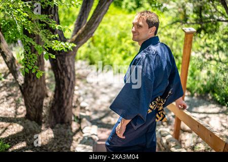 Junger Mann im Kimono-Kostüm, der sich im japanischen Garten mit Blick auf die Natur auf einen Geländerzaun lehnt und den Kopf dreht Stockfoto