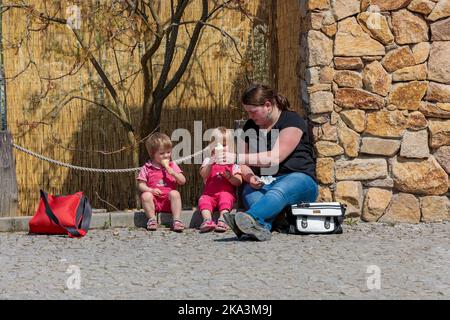 Tschechische Republik Prag April 2019 Eine junge Frau mit zwei Kindern sitzt auf dem Bürgersteig in einem Stadtpark. Mama füttert Kinder Eis. Ruhe nach einem Spaziergang. Stockfoto