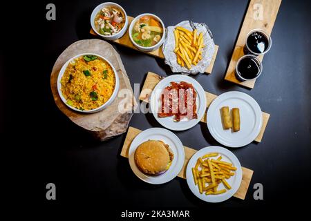 Ein Blick von oben auf gelben Reis, pommes frites, Rippchen, Burger und Meeresfrüchte auf weißen Tellern mit zwei Tassen Limonade, die auf Holzbrettern auf dem Tisch serviert werden Stockfoto