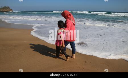 Eine Rückansicht einer Mutter in Heijab und ihrer Tochter, die am Strand entlang läuft und die Meereswellen ins Ufer krachen Stockfoto