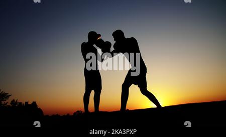 Zwei dunkle männliche Figuren, bei Sonnenaufgang, gegen das Licht, Boxen, im Sparring kämpfen, Training in zwei Streiktechniken. Am Sandstrand, im Frachthafen, am Wasser, im Sommer. Hochwertige Fotos Stockfoto