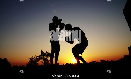 Zwei dunkle männliche Figuren, bei Sonnenaufgang, gegen das Licht, Boxen, im Sparring kämpfen, Training in zwei Streiktechniken. Am Sandstrand, im Frachthafen, am Wasser, im Sommer. Hochwertige Fotos Stockfoto