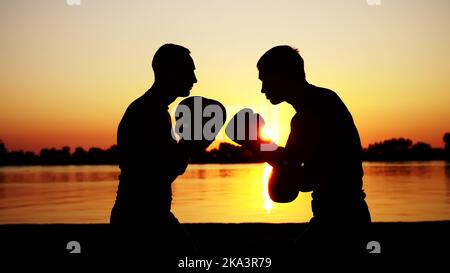 Zwei dunkle männliche Figuren, bei Sonnenaufgang, gegen das Licht, Boxen, im Sparring kämpfen, Training in zwei Streiktechniken. Am Sandstrand, im Frachthafen, am Wasser, im Sommer. Hochwertige Fotos Stockfoto