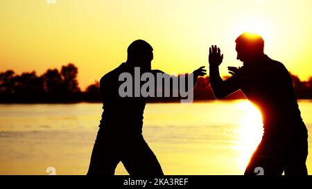 Zwei dunkle männliche Figuren, bei Sonnenaufgang, gegen das Licht, Boxen, im Sparring kämpfen, Training in zwei Streiktechniken. Am Sandstrand, im Frachthafen, am Wasser, im Sommer. Hochwertige Fotos Stockfoto