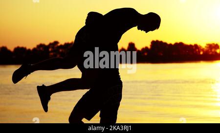 Zwei dunkle männliche Figuren, bei Sonnenaufgang, gegen das Licht, Boxen, im Sparring kämpfen, Training in zwei Streiktechniken. Am Sandstrand, im Frachthafen, am Wasser, im Sommer. Hochwertige Fotos Stockfoto