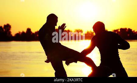 Zwei dunkle männliche Figuren, bei Sonnenaufgang, gegen das Licht, Boxen, im Sparring kämpfen, Training in zwei Streiktechniken. Am Sandstrand, im Frachthafen, am Wasser, im Sommer. Hochwertige Fotos Stockfoto