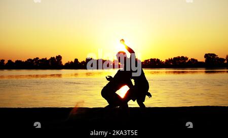 Zwei dunkle männliche Figuren, bei Sonnenaufgang, gegen das Licht, Boxen, im Sparring kämpfen, Training in zwei Streiktechniken. Am Sandstrand, im Frachthafen, am Wasser, im Sommer. Hochwertige Fotos Stockfoto