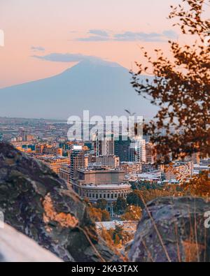 Ein vertikaler atemberaubender Blick auf das Stadtbild von Jerewan hinter den Felsen mit der Silhouette des Berges Ararat im Hintergrund Stockfoto