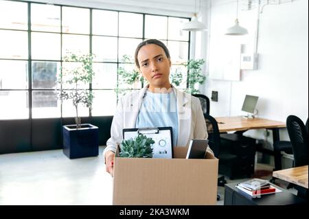 Job verloren. Unglückliche, enttäuschte Latino- oder brasilianerin verärgert, steht in ihrem Arbeitsbüro, hält einen Karton mit Arbeitsattributen in den Händen, schaut traurig auf die Kamera, hat ihren Job verloren, gefeuert Stockfoto