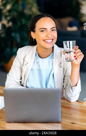 Vertikales Foto einer glücklichen, erfolgreichen, gemischten Arbeiterin, die ein Glas Mineralwasser hält, während sie am Arbeitsplatz im Büro sitzt, sich gesund fühlt, sich um die Gesundheit kümmert und die Kamera lächelt Stockfoto