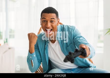 Zufrieden aufgeregt jungen afroamerikanischen Mann mit Joystick spielen Online-Spiel, freut sich zu gewinnen, sitzt auf dem Stuhl Stockfoto