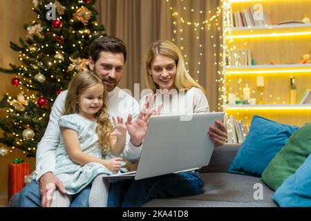 Glückliche Familie begrüßt Freunde per Videochat, Ehemann, Ehefrau und Tochter sitzen zu Hause auf dem Sofa und winken die Hände, Online-Fernkommunikation und Grüße zu Weihnachten und Neujahr. Stockfoto