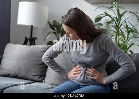 Ungesundes junges Mädchen, das auf dem Sofa sitzt, leidet an Menstruationskrämpfen, verärgert unglückliche junge Brünette, trauert, leidet nach Fehlgeburten oder Abort Stockfoto