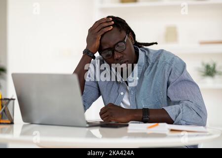 Müder Schwarzer Mann, Der Am Schreibtisch Sitzt, Mit Laptop Und Berührtem Kopf Stockfoto