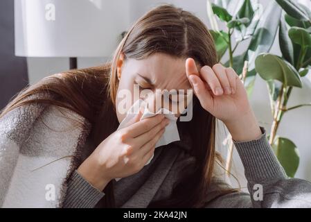 Konzept des geschwächten Immunsystems. Nahaufnahme einer kranken Frau, die auf der Couch im Wohnzimmer sitzt und mit karierten Niesen bedeckt ist und Papierservietten in der Hand hält Stockfoto