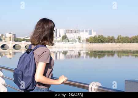 Eine Rückansicht eines jungen Studenten mit lockigen Haaren, der in einem T-Shirt und Jeans und einem blauen Rucksack vor dem Hintergrund in einem Park steht Stockfoto