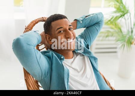Fröhlicher, gutaussehender, tausendjähriger afroamerikanischer Mann, der allein auf einem Stuhl ruht, genießt Komfort und Ruhe Stockfoto