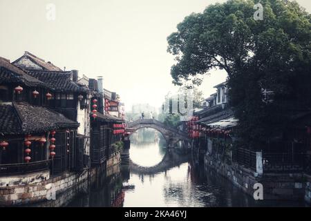 Eine malerische Ansicht einer gewölbten Brücke über einen Kanal mit Reflexion auf dem Wasser und Gebäuden mit roten traditionellen chinesischen Laternen in Wuzhen, China Stockfoto