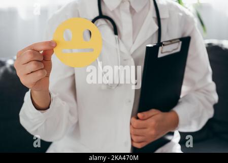 Nahaufnahme einer Ärztin in weißer Uniform mit Stethoskop, das ein gelbes trauriges Gesicht mit Papier hält, und einem schlechten Smiley, der auf dem Sofa sitzt. Feedback-Bewertung und Kundenrev Stockfoto