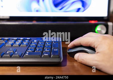 Selektiver Fokus auf eine Hand, die auf eine Maus klickt, während Monitor und Soundbar im Hintergrund unscharf sind. Stockfoto