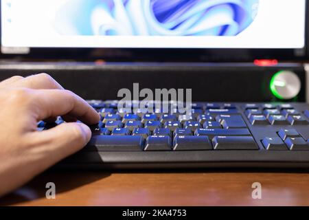 Selektiver Fokus auf eine Hand, die auf einer Tastatur tippt, während Monitor und Soundbar im Hintergrund unscharf sind. Stockfoto