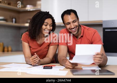 Glückliche junge schwarze Dame im roten T-Shirt zeigt dem Mann mit Computer im Kücheninnenraum Dokumente Stockfoto