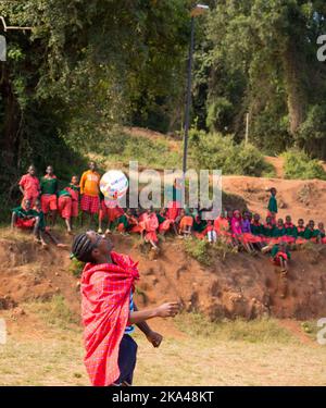 Eine vertikale Aufnahme eines jungen Mannes mit einem eingewickelten roten Umhang, der vor Kindern mit einem Ball Tricks macht. Loita Hills, Kenia. Stockfoto
