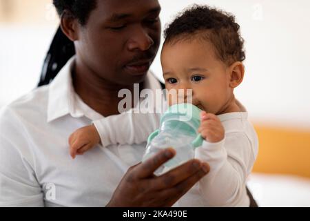 Fürsorglicher Afroamerikanischer Vater, Der Seinem Niedlichen Kleinen Baby Wasserflasche Gibt Stockfoto