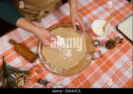 Details: Hände einer Frau Hausfrau, die Mehl auf gerollten Teig streut, während sie Lebkuchenkekse für Weihnachten macht Stockfoto