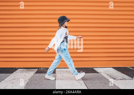 Kleine Mode-gekleidete Mädchen lustig zu Fuß über einen Fußgängerzebra Crosswalk auf der orangefarbenen Wand Hintergrund. Urbane Menschen leben und eine Straße jeden Stockfoto