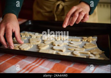 Details: Die Hände einer Hausfrau legten aus Lebkuchenteig geschnittene Kekse auf das Backblech und bereiteten Weihnachtsgebäck zu Stockfoto