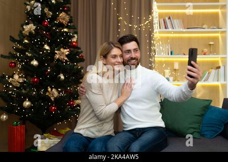 Verheiratetes Paar Mann und Frau zusammen auf dem Sofa zu Hause Weihnachtsbaum mit Smartphone Videoanruf reden und Selfie, reife Familie Gruß Freunde online für Neujahr und Weihnachten. Stockfoto