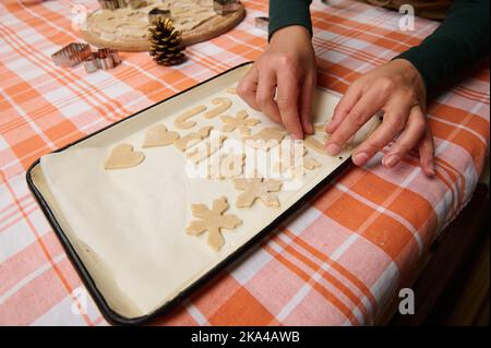 Die Hände der Frau, die Konditorin auslagend, schneiden die Formen aus dem Lebkuchenteig, auf dem Backblech aus, für die Platzierung in den Ofen Stockfoto