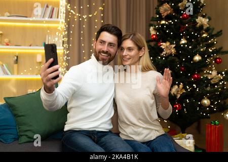 Verheiratetes Paar Mann und Frau zusammen auf dem Sofa zu Hause Weihnachtsbaum mit Smartphone Videoanruf reden und Selfie, reife Familie Gruß Freunde online für Neujahr und Weihnachten. Stockfoto