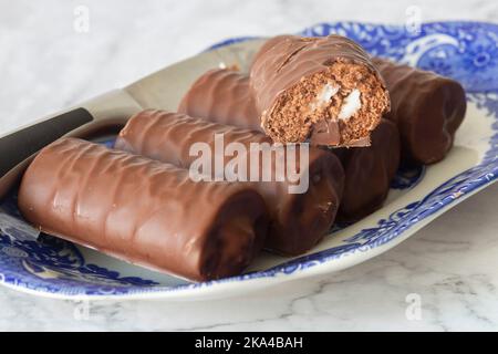 Einzelne Schokoladenschwammrollen auf einer gemusterten blau-weißen Schale mit einem Serviermesser. Auf weißem Marmorboden Stockfoto