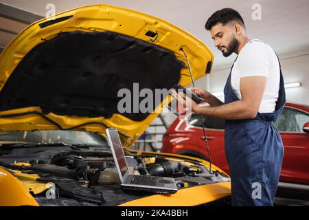 Serious fokussierten Mann Auto Techniker Mechaniker Reparatur Auto Problem des Motors, während der Systemprüfung Detail, mit Tablet-Computer für die Wartung und Befestigung in der Garage. Stockfoto