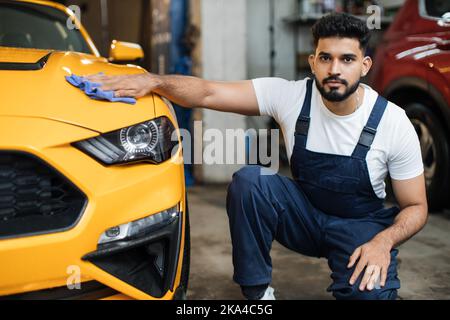Bärtiger Autowascharbeiter, trägt Schutzkleidung Uniform Overalls, Reinigung modernen gelben Luxus-Auto mit Mikrofaser Tuch. Fahrzeugdetails oder Fahrzeugkonzept. Stockfoto