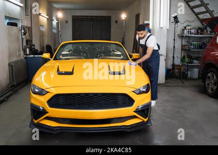 In voller Länge Porträt von Auto waschen Arbeiter Mann, trägt Schutzkleidung Uniform Overalls, Reinigung moderne gelbe Luxus-Auto mit Mikrofaser Tuch. Fahrzeugdetails oder Fahrzeugkonzept. Stockfoto