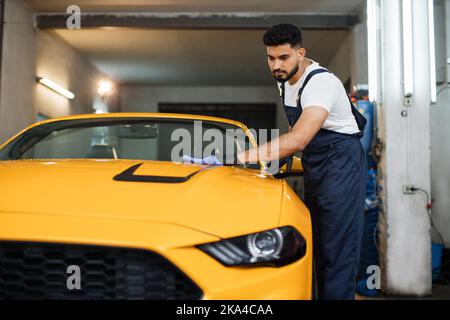 In voller Länge Porträt von Auto waschen Arbeiter Mann, trägt Schutzkleidung Uniform Overalls, Reinigung moderne gelbe Luxus-Auto mit Mikrofaser Tuch. Fahrzeugdetails oder Fahrzeugkonzept. Stockfoto