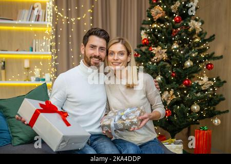 Porträt eines erwachsenen bärtigen Mannes und einer blonden Frau, die die Kamera in der Nähe des Weihnachtsbaums betrachten, und eines Paares, das Weihnachten und Neujahr mit Geschenken zu Hause auf dem Sofa im Wohnzimmer feiert. Stockfoto