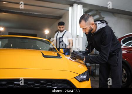 Team von jungen bärtigen Männern, Arbeiter des Auto-Detaillierungsservice. Männer hält Orbital Polisher in der Hand und poliert Haube und wischen Windschutzscheibe des gelben modernen Autos. Auto Detaillierung und Polieren Konzept. Stockfoto